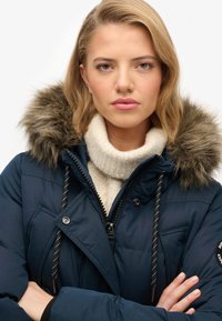 Donkerblauwe pufferjack met een nepbontkap, een rits aan de voorkant en zwart-witte trekkoorden, over een crème gebreide coltrui.