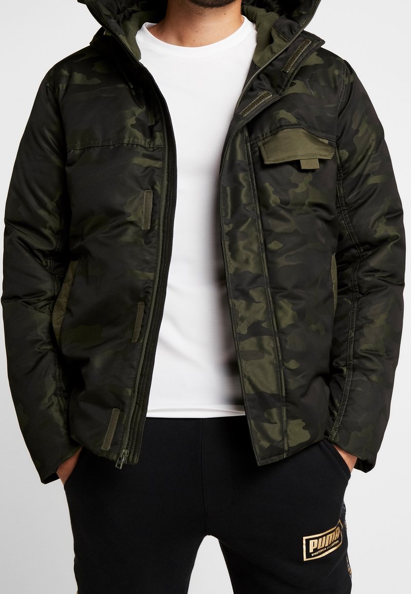 Czarna zielona kurtka w kamuflażu z zapięciem na zamek, kapturem i kieszenią na piersi; ma pikowaną teksturę i elastyczne mankiety. Noszona na białej koszuli.