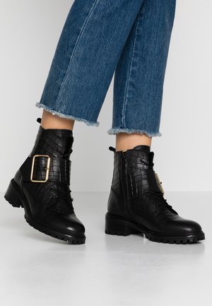 Botas de tobillo de cuero negro texturizado con un gran hebilla dorada y cremallera lateral, combinadas con jeans azul desgastados en un fondo de suelo claro.