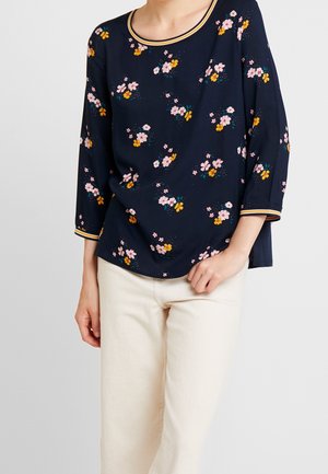 Kvinde iført navyblå bluse med lange ærmer med pink og gult blomsterprint og stribet kant, kombineret med off-white bukser.