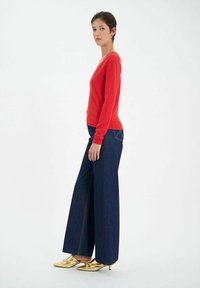 Pull rouge à col en V, associé à un jean en denim bleu foncé à jambes larges. Le mannequin porte des escarpins dorés à bout pointu. Design simple et textures lisses.