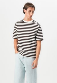Ung mann som har på seg en svart og hvit stripete t-skjorte og lyseblå jeans, står med en hånd i lommen og ser nedover til siden.