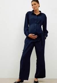 Marineblaue Umstandsmode mit einer geknöpften Bluse und einem Bindeband in der Taille sowie lockeren, geraden Hosen aus weichem Stoff.