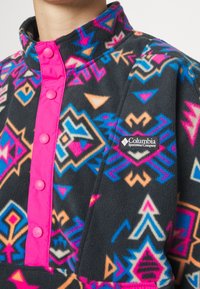 Fleece-pullari, jossa on musta pohja ja värikkäitä geometrisia kuvioita vaaleanpunaisena, sinisenä ja oranssina. Edessä nappikiinnitys ja logotwiisti.