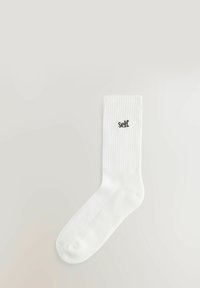 Biela bavlnená ponožka s krátkym strihom a rebrovanou textúrou, s hnedým vyšitým logom "self" blízko lemu. Klasický dizajn s jednofarebným prevedením.