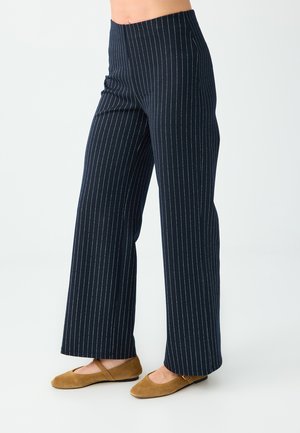 Femme portant un pantalon large taille haute bleu marine à fines rayures et des chaussures plates en daim marron, debout sur un fond blanc.