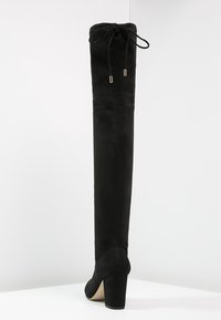 Bota negra de ante por encima de la rodilla con puntera redondeada y tacón alto de bloque. Presenta un diseño con cordones en la parte superior y una textura suave.
