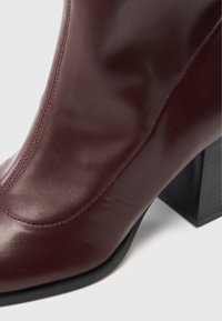 Bottine en cuir bordeaux avec une finition lisse, dotée d'un bout carré et d'un talon noir épais. Des coutures visibles soulignent le design.