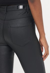 Zwarte, aansluitende jeans van een slick materiaal, met een achterzak met subtiele stiksel en een glanzend merkembleem op de tailleband.