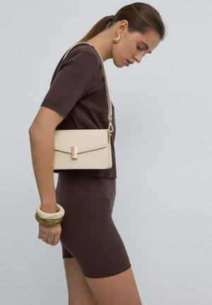 Femme portant un haut marron à manches courtes et une jupe, avec des boucles d'oreilles dorées, tenant un sac bandoulière beige et deux bracelets épais sur fond gris.