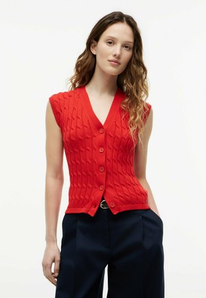Giovane donna con capelli mossi che indossa un gilet rosso lavorato a maglia senza maniche con bottoni e pantaloni scuri a vita alta, in piedi con una mano in tasca.