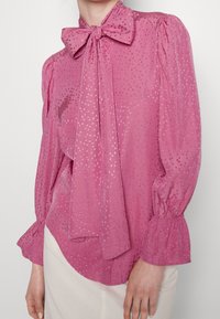 Rosa blus med en knytning vid halsen, puffärmar och ett strukturerat mönster med små, metalliska accenter. Lätt, flödande tyg.