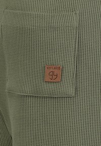 Groene wafelbreisel stof met een vierkante zak voorzien van een bruine leren patch met het label "KEY LARGO" en een gestileerd logo.