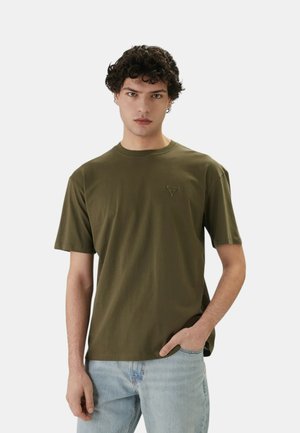 Giovane uomo con capelli ricci, che indossa una maglietta verde oliva a maniche corte e jeans azzurri, in piedi contro uno sfondo semplice.