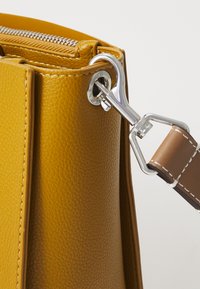 Borsa di colore giallo senape in pelle testurizzata, con dettagli in metallo argentato e una tracolla marrone con cuciture bianche. Chiusura con zip visibile nella parte superiore.