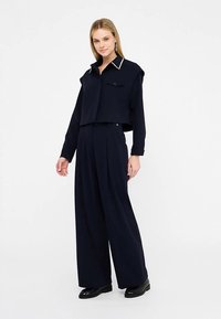 Mornarsko plavi dvodijelni komplet koji uključuje kratku košulju s kopčanjem i šiljastim ovratnikom te odgovarajuće široke hlače visokog struka. Glatki materijal.