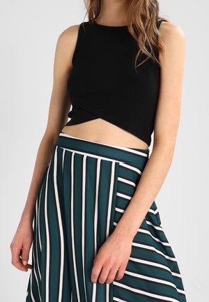 Femme portant un crop top noir sans manches et une jupe teal taille haute avec des rayures verticales blanches et noires.