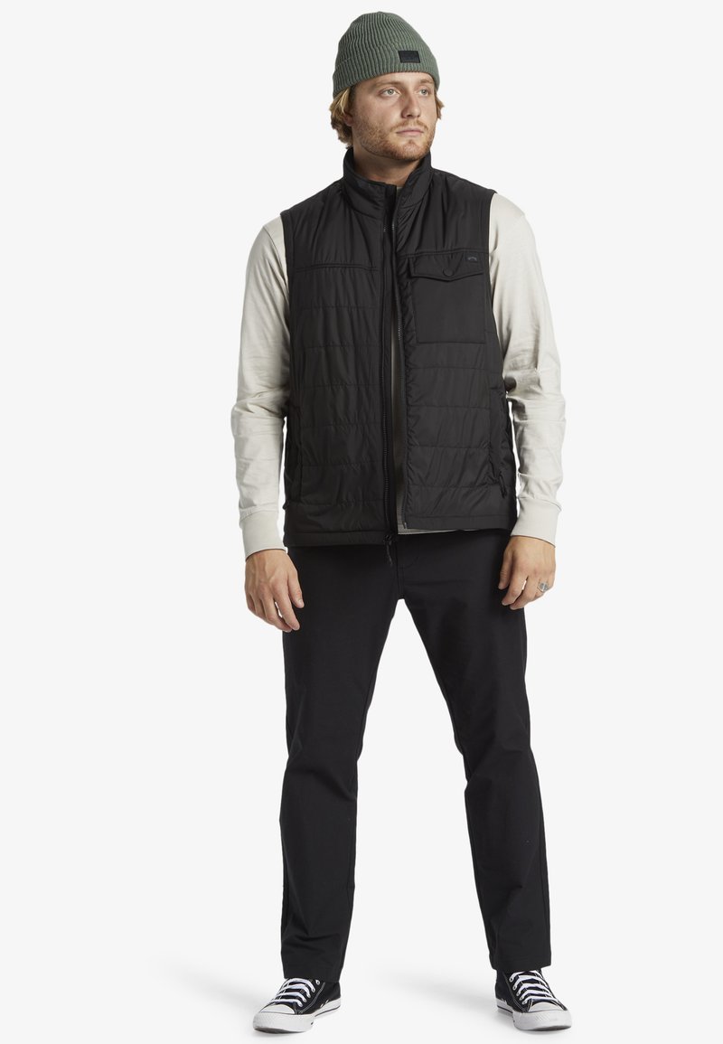 Sort puffer vest med lynlås foran og en brystlomme, der bæres over en lys grå langærmet skjorte. Sorte bukser og sorte sneakers fuldender looket.