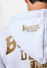 Felpa bianca con grande stampa dorata con la scritta "BOXEUR DES RUES." Texture morbida con un design centralizzato; cappuccio e dettagli posteriori visibili.