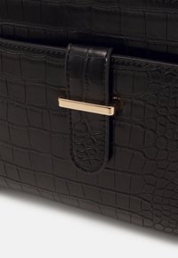 Czarna torba z ekoskóry o fakturze wzoru krokodyla, z prostokątnym zapięciem z metalu w złotym odcieniu i przeszyciami jako akcentami.