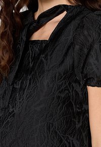 Femme portant une robe noire texturée avec un décolleté carré et un détail découpé près de la clavicule, dotée de manches courtes bouffantes.