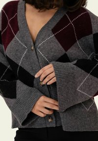 Grauer Cardigan mit Rautenmuster in Weinrot, Schwarz und Weiß, mit gerippten Bündchen und zwei Knöpfen vorne. Weiches Strickmaterial.