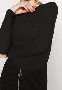 Musta ribbed pitkähiha päällyspaita, jossa on pyöreä kaula-aukko. Kangas on tekstuurikuvioinen; yhdistettynä vetoketjulliseen mustaan hameeseen, jossa on sormivetoketju.