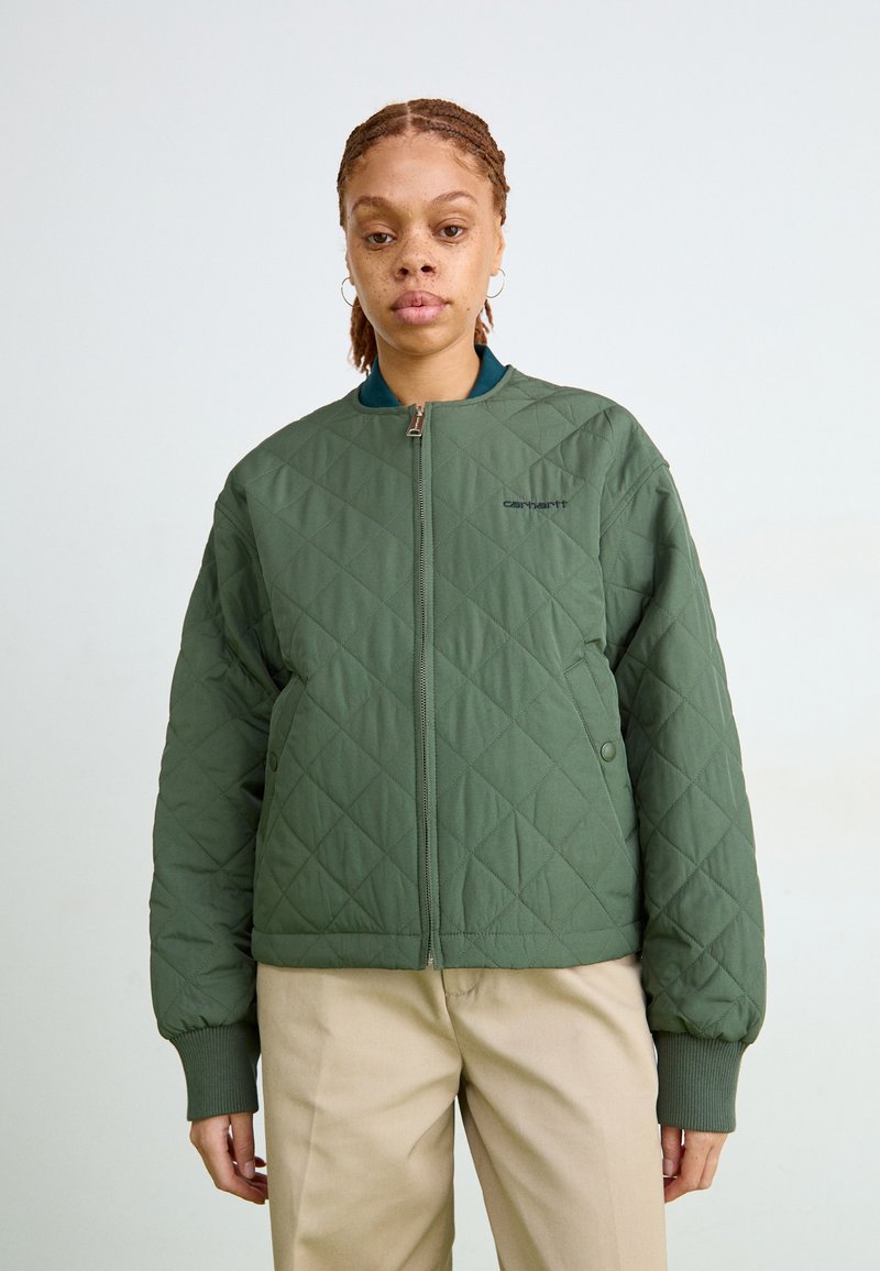 Prešívaná zelená bomber bunda so zipsom na prednej strane, rebrovanými manžetami a malým logom. Nosená na tyrkysovom topu a khaki nohaviciach. Hladká textúra.