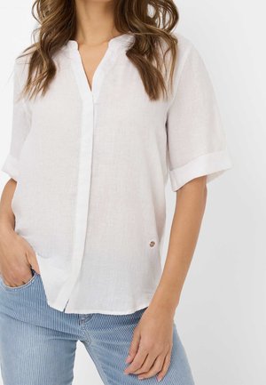 Femme portant un chemisier blanc ample à manches courtes avec des revers roulés et un pantalon bleu rayé, une main dans la poche, sur fond uni.