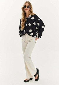 Czarna bluzka z długim rękawem w białe kropki, połączona z kremowymi dżinsami w stylu dzwonów i czarnymi klapkami. Okulary przeciwsłoneczne dopełniają strój.