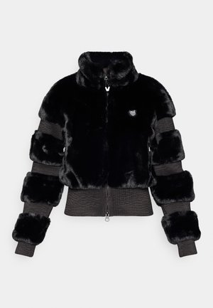 Giacca in pelo sintetico nero con maniche a maglia rigata, colletto alto, chiusura con zip e patch con logo sul petto; presenta una texture morbida e un design cropped.