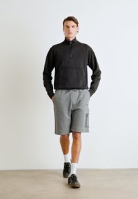 Musta vetoketjullinen pullover etutaskulla, yhdistettynä harmaisiin shortseihin, joissa on sivutasku ja mustat yksityiskohdat. Mallilla on päällään mustat lenkkarit.