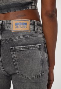 Hallitus hallid teksased, millel on kulunud viimistlus. Omab nahkavat lugedes "MOSCHINO JEANS" sinises kirjas. Tagumise tasku detail on nähtav.