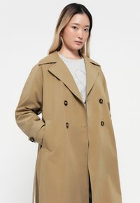Cappotto trench beige doppiopetto con un ampio colletto, grandi bottoni neri e tasche laterali, in contrasto con una camicia grigia sottostante.