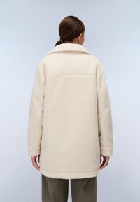 Casaco de cor creme forrado a felpa, com colarinho, design ligeiramente oversized e tecido texturizado, visto de costas.