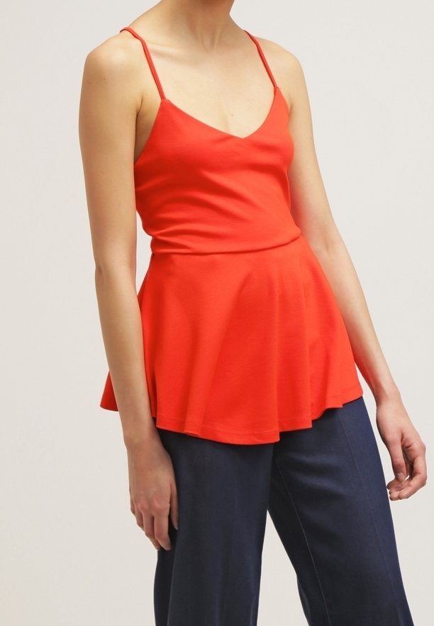 Femme portant un top peplum sans manches rouge vif à fines bretelles, associé à un pantalon bleu foncé, debout devant un fond uni.