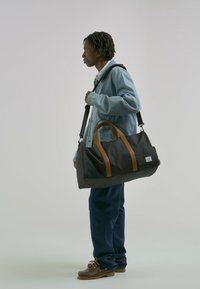 Čierna cestovná taška s hnedými koženými rukoväťami a popruhom, s obdĺžnikovým tvarom, hladkou textúrou a logom na náplasti.