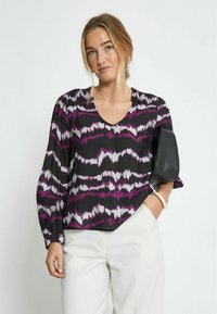Blusa nera con motivo astratto a onde viola e bianche, scollo a v, maniche lunghe e vestibilità ampia, abbinata a pantaloni bianchi e una borsa nera.