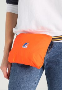 Personne portant un jean bleu et un sweat-shirt blanc avec une sacoche ceinture K-Way orange vif autour de la taille.