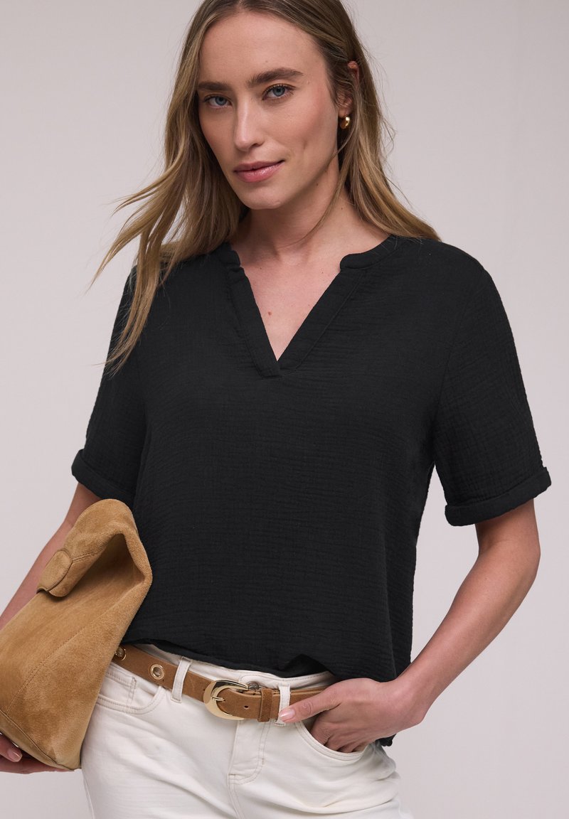 Femme aux longs cheveux blonds portant un haut noir à col en V, un pantalon blanc avec une ceinture beige, tenant une pochette en daim beige.