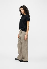 Γυναίκα με σγουρά μαλλιά που φοράει μαύρο κοντομάνικο πουκάμισο, μπεζ καρό παντελόνι με φαρδιά πόδια και μαύρα γυαλιστερά παπούτσια, στέκεται πλαγίως.