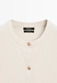 Cardigan in cotone color crema con scollo rotondo e due bottoni decorativi dorati sul davanti. L'etichetta riporta "Massimo Dutti".