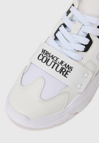 Ténis branco que combina couro e rede, com um design distinto em forma de onda, tira ajustável e marca "VERSACE JEANS COUTURE".