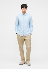 Νεαρός άνδρας στέκεται με τα χέρια στις τσέπες, φορώντας ανοιχτό μπλε πουκάμισο με κουμπιά, μπεζ παντελόνι cargo και μπεζ παπούτσια τύπου slip-on, μπροστά σε λευκό φόντο.