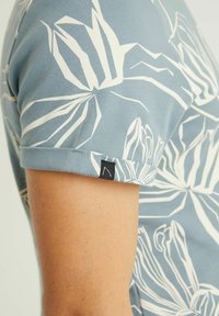 Lyhythihainen paita vaaleansinisestä kankaasta, jossa on valkoinen kukkakuosi ja käännetyt hihat. Paidassa on pieni musta merkki helman kohdalla.