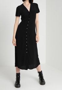 Černé midi šaty s krátkými nafouknutými rukávy, výstřihem do V a zapínáním na knoflíky vpředu. Mají hladkou texturu a sahají těsně pod kolena. Nosit s černými botami.