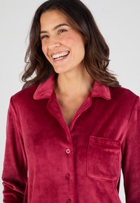 Camisa de pijama de terciopelo rojo con cuello, mangas largas y un bolsillo en el pecho; cuenta con cierre de botones y una textura suave y lisa.