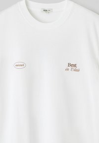T-shirt en coton blanc avec une coupe ample. Présente des éléments brodés : "winner" dans un ovale marron et "Best in Class" en texte marron.