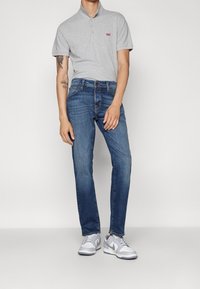 Polo grigia con due bottoni, jeans di denim blu e scarpe da ginnastica grigie con dettagli bianchi. Vestibilità aderente e design casual.