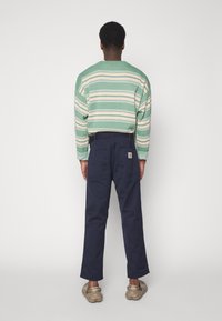 Sweter w paski z zielonym i beżowym splotem, zestawiony z ciemnogranatowymi spodniami. Teksturowana tkanina o swobodnym kroju, z dwoma tylnymi kieszeniami.